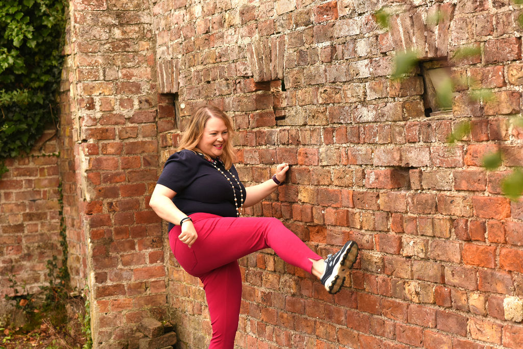 Picture of Claire Bradley, a blonde haired woman wearing pink trousers and a navy top kicking a brick wall